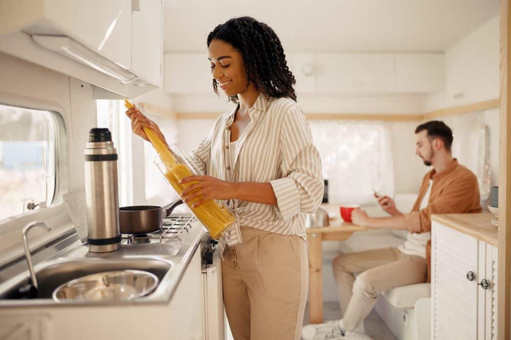 Woman holds a serving of dry pasta noodles in her hands, as her partner sits at the RV table in the background.